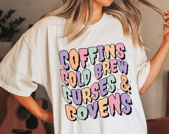 Op de afbeelding: Een wit oversized T-shirt met een kleurrijke grafische print met de tekst "Coffins Cold Brew Curses & Covens".