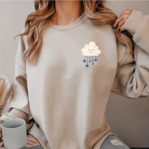 May include: A light gray sweatshirt with a white cloud graphic with a smiling face and blue raindrops. The cloud has a stitched design and the raindrops are hearts.
