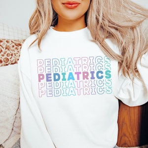 A model wears a white sweatshirt that says Pediatrics. It is written 5 times repeated. Middle line is solid letter, outer 4 are outlined letter. Each letter is different color: muted mauve, muted, purple, darker blue, light teal, med-blue, pink.
