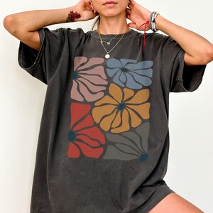 A model wears a pepper shirt with abstract, wavy, boho flowers in colors of muted mauve, light grey-blue, gold, red, and dark taupe all with deep blue pistils. The colorful design is a slightly distressed texture.