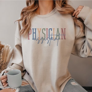 May include: A light beige sweatshirt with the text "PHYSICIAN assistant" printed in colorful letters. The text is in a rainbow color scheme with a light blue background.