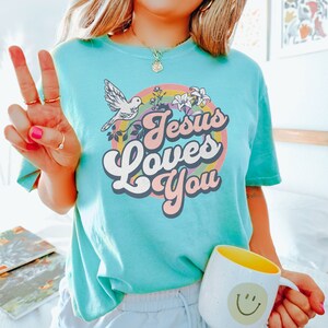 May include: A turquoise blue t-shirt with a retro rainbow design and the text "Jesus Loves You".