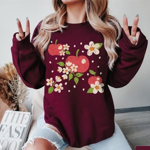 A model wears a maroon sweatshirt with a retro inspired and slightly distressed graphic of 3 red apples surrounded by delicate cream flowers and some cream colored apple seeds around.