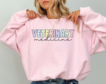 Op de afbeelding: Een lichtroze sweatshirt met de tekst "VETERINARY medicine" in regenboogkleuren.