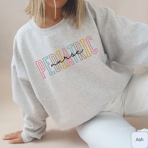 A model wears ash  sweatshirt that says Pediatric nurse. Pediatric is in outlined blocky font. Each letter a different color: melon-pink, med-bright-blue, dark taupe, yellow-gold. Nurse is smaller lowercase script in the middle.