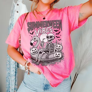 May include: Pink t-shirt with the text "Summerween Vibes" and a graphic of a skeleton relaxing on a flamingo floatie, surrounded by jack-o'-lanterns and bats. The shirt is a vibrant pink color.