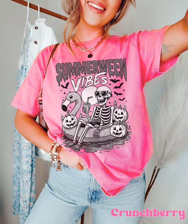 May include: Pink t-shirt with the text "Summerween Vibes" and a graphic of a skeleton relaxing on a flamingo floatie, surrounded by jack-o'-lanterns and bats. The shirt is a vibrant pink color.