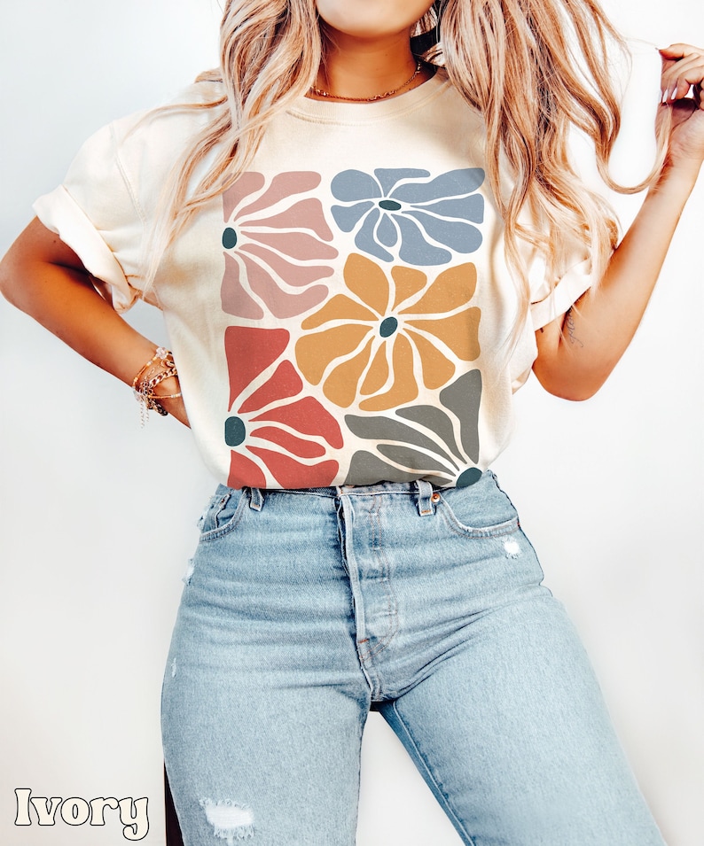 A model wears an ivory shirt with abstract, wavy, boho flowers in colors of muted mauve, light grey-blue, gold, red, and dark taupe all with deep blue pistils. The colorful design is a slightly distressed texture.