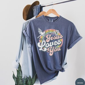 May include: A navy blue t-shirt with a rainbow and a dove graphic. The text "Jesus Loves You" is in a retro font.