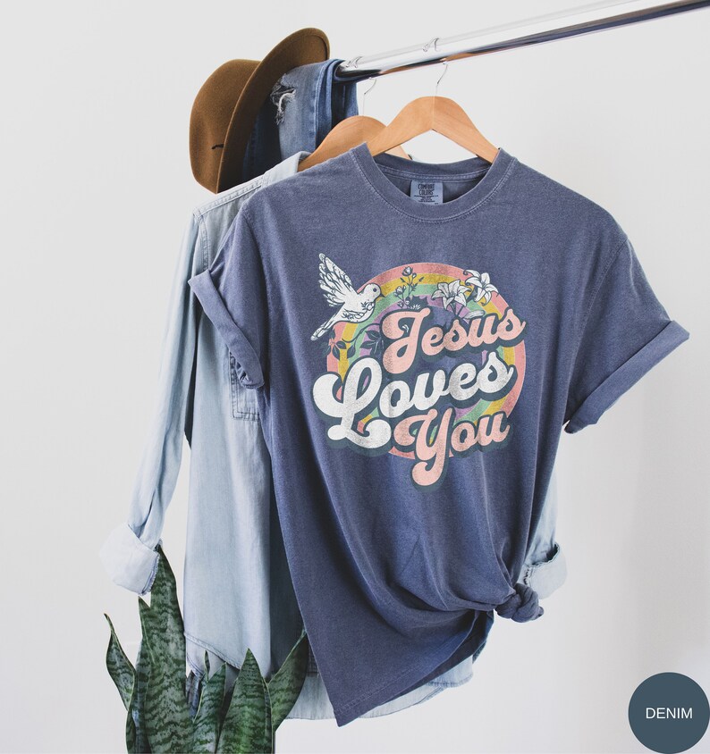 May include: A navy blue t-shirt with a rainbow and a dove graphic. The text "Jesus Loves You" is in a retro font.