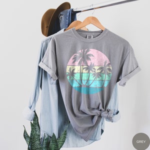以下が含まれることがあります： グレーのTシャツに、ピンク、イエロー、グリーン、ブルーのヴィンテージスタイルのヤシの木と夕焼けのグラフィックがプリントされています。画像の右下には「GREY」というテキストが表示されています。