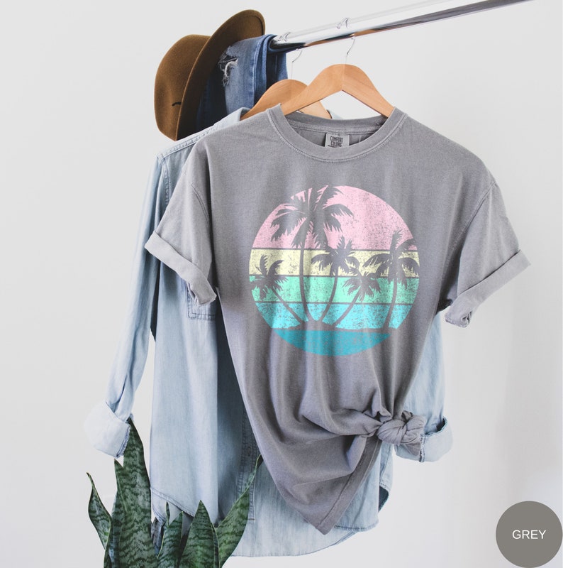 以下が含まれることがあります： グレーのTシャツに、ピンク、イエロー、グリーン、ブルーのヴィンテージスタイルのヤシの木と夕焼けのグラフィックがプリントされています。画像の右下には「GREY」というテキストが表示されています。