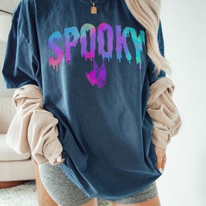 May include: Midnight blue t-shirt with the word "SPOOKY" in a colorful, dripping font. A bat graphic is below the word. The shirt is part of a Halloween-themed collection.