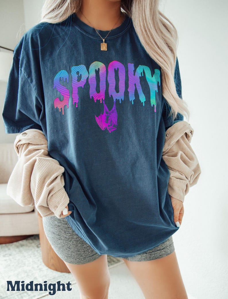 May include: Midnight blue t-shirt with the word "SPOOKY" in a colorful, dripping font. A bat graphic is below the word. The shirt is part of a Halloween-themed collection.