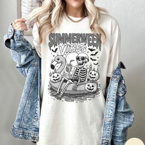 May include: Ivory-colored t-shirt with a graphic featuring a skeleton in sunglasses relaxing on a flamingo floatie, surrounded by jack-o'-lanterns and bats. The text "Summerween Vibes" is above the graphic. The shirt is worn with a denim jacket.