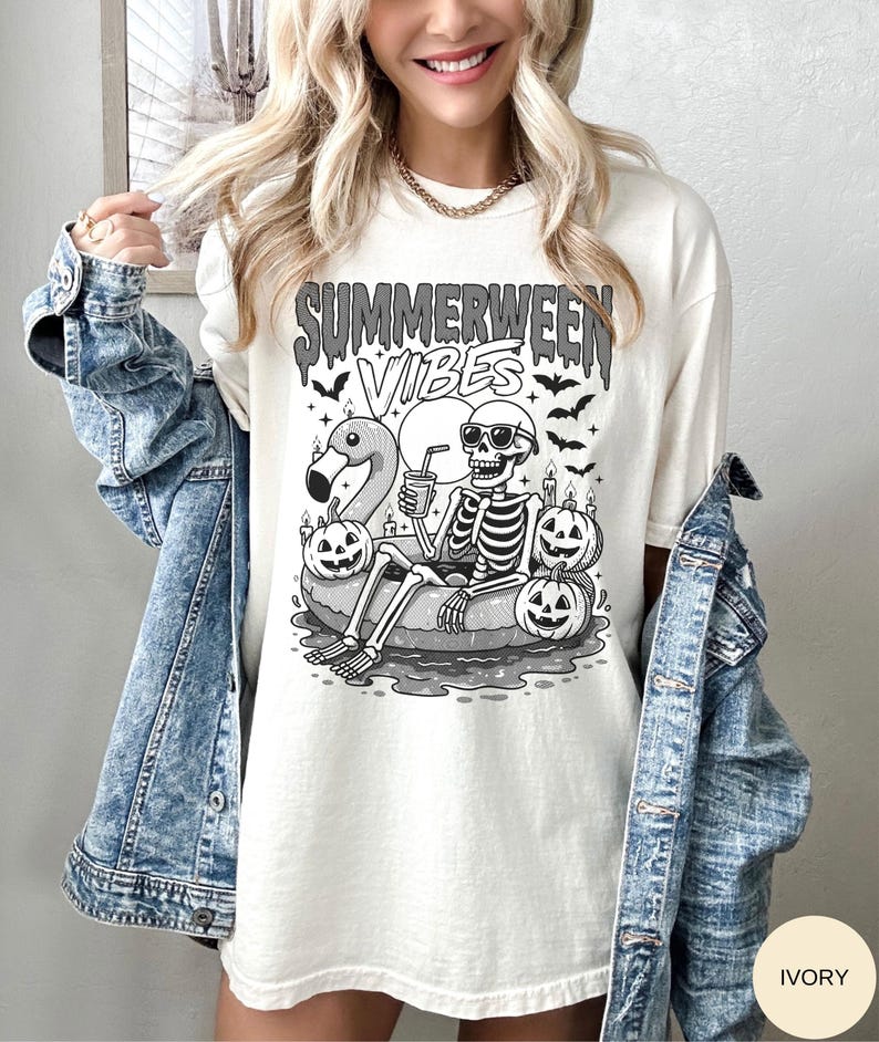 May include: Ivory-colored t-shirt with a graphic featuring a skeleton in sunglasses relaxing on a flamingo floatie, surrounded by jack-o'-lanterns and bats. The text "Summerween Vibes" is above the graphic. The shirt is worn with a denim jacket.