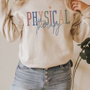 May include: A light beige sweatshirt with the text "PHYSICAL therapy" in colorful letters. The text is in a rainbow color scheme with a blue, red, orange, and green color palette. The sweatshirt is being worn by a person with long blonde hair. The person is wearing blue jeans.