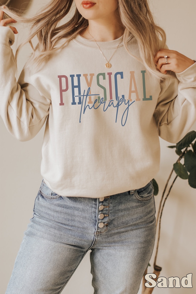 May include: A light beige sweatshirt with the text "PHYSICAL therapy" in colorful letters. The text is in a rainbow color scheme with a blue, red, orange, and green color palette. The sweatshirt is being worn by a person with long blonde hair. The person is wearing blue jeans.