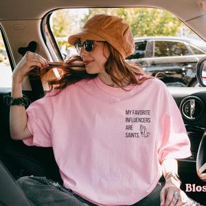 May include: A pale pink t-shirt featuring the text "MY FAVORITE INFLUENCERS ARE SAINTS" and a praying hands graphic. The shirt is worn by a person in a car, with a corduroy hat and sunglasses. The word "Blossom" is in the bottom right corner.