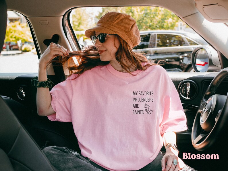 May include: A pale pink t-shirt featuring the text "MY FAVORITE INFLUENCERS ARE SAINTS" and a praying hands graphic. The shirt is worn by a person in a car, with a corduroy hat and sunglasses. The word "Blossom" is in the bottom right corner.