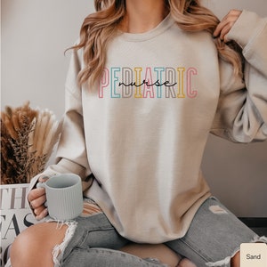 May include: A light beige sweatshirt with the text "PEDIATRIC nurse" in colorful outlined letters. The sweatshirt is worn by a person sitting on a chair with a light blue mug in their hand.