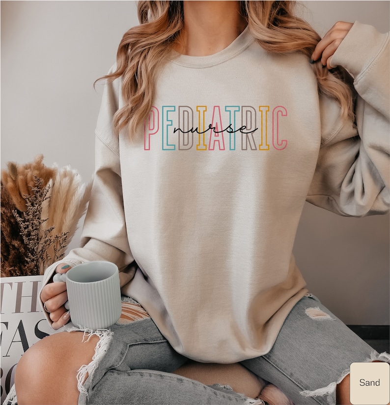May include: A light beige sweatshirt with the text "PEDIATRIC nurse" in colorful outlined letters. The sweatshirt is worn by a person sitting on a chair with a light blue mug in their hand.