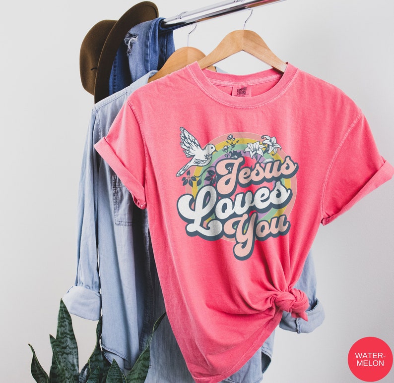 May include: A pink t-shirt with a rainbow and a dove graphic. The text "Jesus Loves You" is in a retro font.