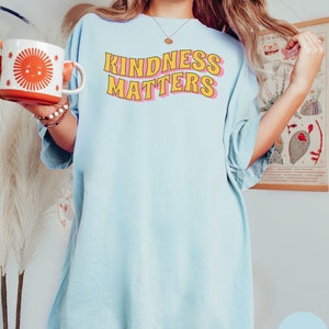 Puede incluir: Una camiseta de gran tama&ntilde;o de color azul claro con el texto "KINDNESS MATTERS" en letras amarillas y rosas.