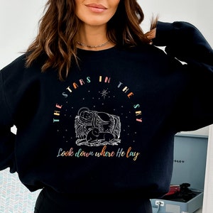 May include: Black sweatshirt with a colorful graphic design featuring a star-shaped circle with the text "The stars in the sky Look down where He lay".