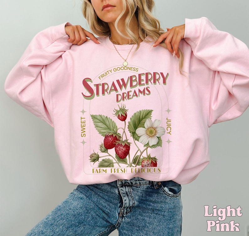 A model wears a light pink sweatshirt with a vintage-inspired graphic of strawberries on the stem. It says Strawberry dreams in an art-deco font with other highlighted words around that say fruity goodness, sweet, juicy, farm fresh, and delicious.