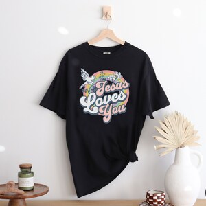May include: Black t-shirt with a retro-style graphic that says "Jesus Loves You" in a rainbow circle with a white dove flying above.
