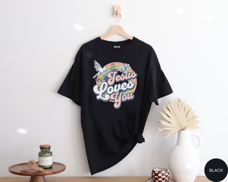 May include: Black t-shirt with a retro-style graphic that says "Jesus Loves You" in a rainbow circle with a white dove flying above.