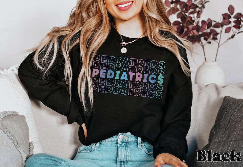 K&ouml;nnte beinhalten: Ein schwarzes Sweatshirt mit dem Wort "PEDIATRICS", das in einem Regenbogen-Farbverlauf wiederholt wird. Das Sweatshirt wird von einer Person getragen, die auf einem Sofa sitzt.