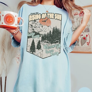 May include: Light blue t-shirt featuring a vintage-style graphic of a mountain landscape and a sunrise. The graphic includes the text "From the Rising of the Sun" and "The name of the Lord is to be praised, Psalm 113:3".
