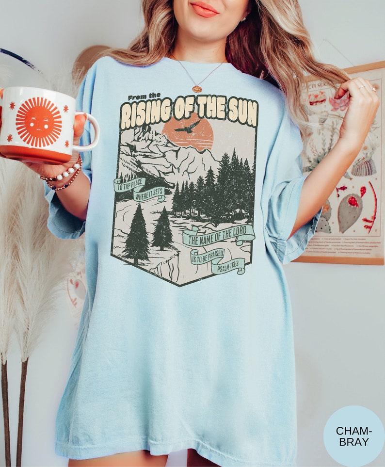 May include: Light blue t-shirt featuring a vintage-style graphic of a mountain landscape and a sunrise. The graphic includes the text "From the Rising of the Sun" and "The name of the Lord is to be praised, Psalm 113:3".