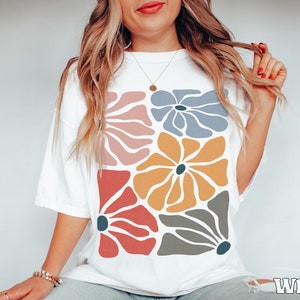 A model wears a white shirt with abstract, wavy, boho flowers in colors of muted mauve, light grey-blue, gold, red, and dark taupe all with deep blue pistils. The colorful design is a slightly distressed texture.