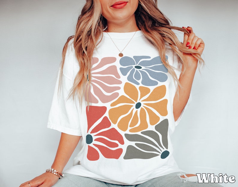 A model wears a white shirt with abstract, wavy, boho flowers in colors of muted mauve, light grey-blue, gold, red, and dark taupe all with deep blue pistils. The colorful design is a slightly distressed texture.