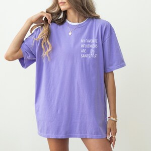 May include: A violet T-shirt featuring the text "MY FAVORITE INFLUENCERS ARE SAINTS" and a graphic of praying hands. The shirt has short sleeves and a loose fit. The model is wearing a necklace and bracelets.
