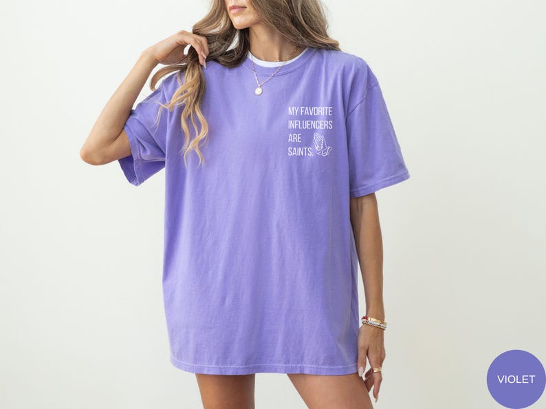 May include: A violet T-shirt featuring the text "MY FAVORITE INFLUENCERS ARE SAINTS" and a graphic of praying hands. The shirt has short sleeves and a loose fit. The model is wearing a necklace and bracelets.