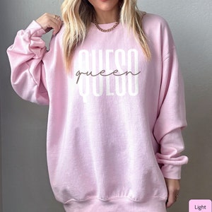 May include: Light pink sweatshirt with the text "queso queen" in white lettering.