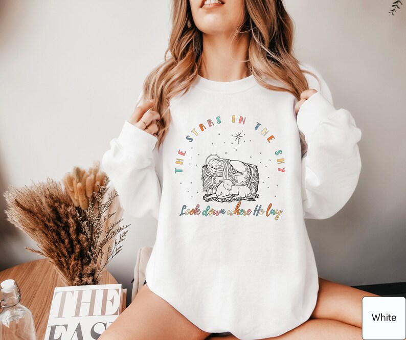 May include: White sweatshirt with a colorful graphic design featuring a manger scene with the text "The stars in the sky Look down where He lay".