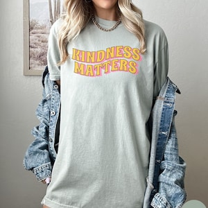 Puede incluir: Una camiseta verde claro con el texto "KINDNESS MATTERS" en letras amarillas y rosas. Las letras est&aacute;n en una fuente de estilo retro.
