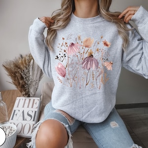 May include: A gray sweatshirt with a floral print featuring pink, brown, and white flowers. The flowers are arranged in a cluster and are surrounded by green leaves.