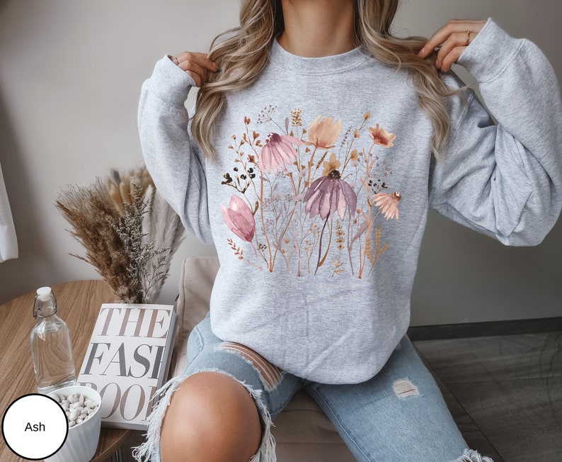 May include: A gray sweatshirt with a floral print featuring pink, brown, and white flowers. The flowers are arranged in a cluster and are surrounded by green leaves.