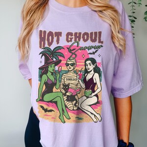 May include: A lilac t-shirt with the text "HOT GHOUL SUMMER" in a retro style. The graphic shows three cartoon ghouls in swimwear on a beach: a witch, a mummy, and a vampire. The mummy wears sunglasses and the word "SUNBLOCK" is on the bandages.