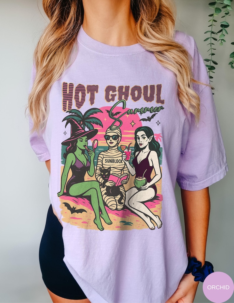 May include: A lilac t-shirt with the text "HOT GHOUL SUMMER" in a retro style. The graphic shows three cartoon ghouls in swimwear on a beach: a witch, a mummy, and a vampire. The mummy wears sunglasses and the word "SUNBLOCK" is on the bandages.
