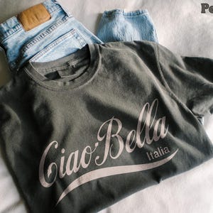 A pepper shirt is folded and says ciao-bella is a classic script style. It has a wavy ribbon underneath and says Italia.