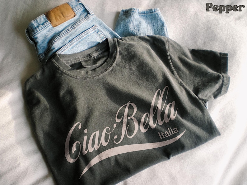 A pepper shirt is folded and says ciao-bella is a classic script style. It has a wavy ribbon underneath and says Italia.