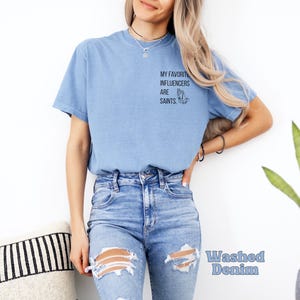 May include: Pale blue t-shirt featuring the text "MY FAVORITE INFLUENCERS ARE SAINTS" and a praying hands graphic. The shirt is made of a soft material and is labelled "Washed Denim".