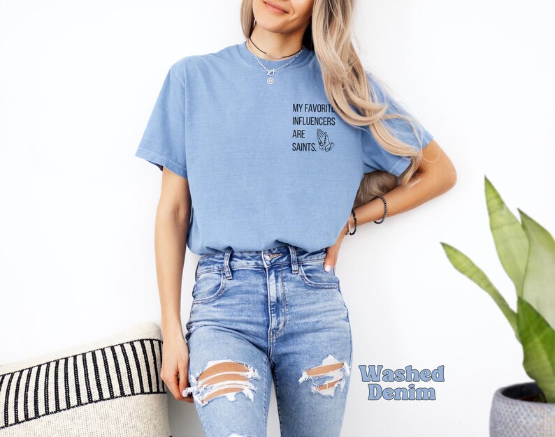 May include: Pale blue t-shirt featuring the text "MY FAVORITE INFLUENCERS ARE SAINTS" and a praying hands graphic. The shirt is made of a soft material and is labelled "Washed Denim".